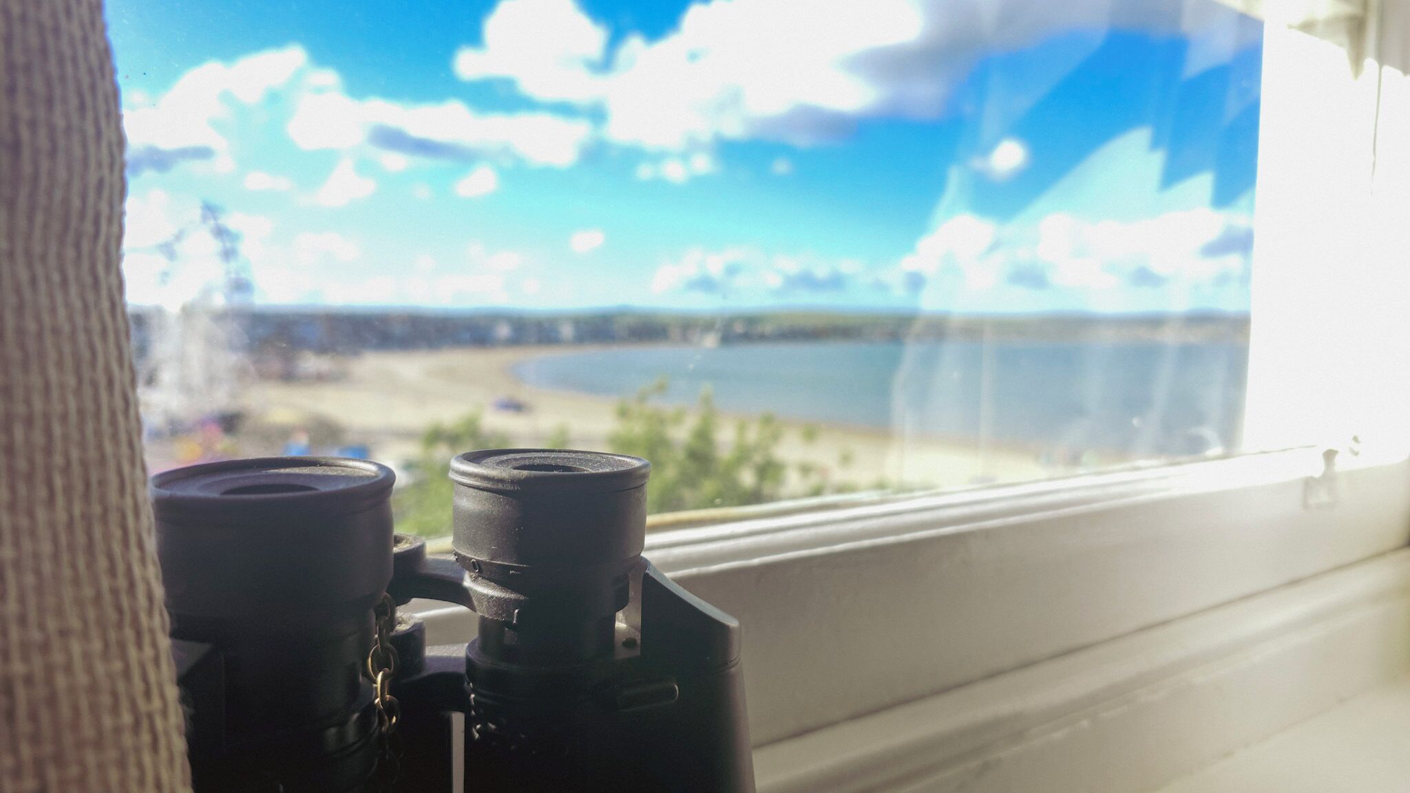 A telescope on a window sill with a view of a beach and water.