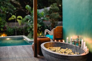 A wooden deck with a stone basin filled with water and floating petals, surrounded by candles and a wooden chair.