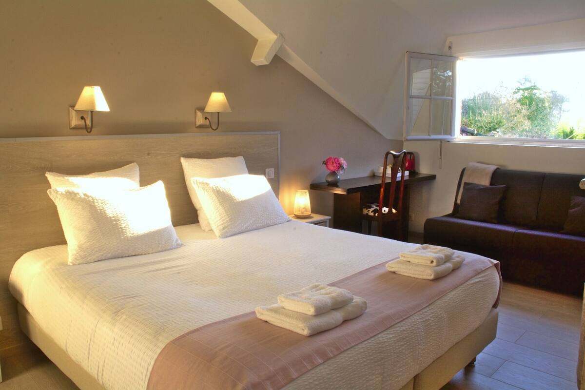 A bedroom with a large bed, two bedside lamps, a nightstand with a lamp, a dresser, and a window with a view of greenery.