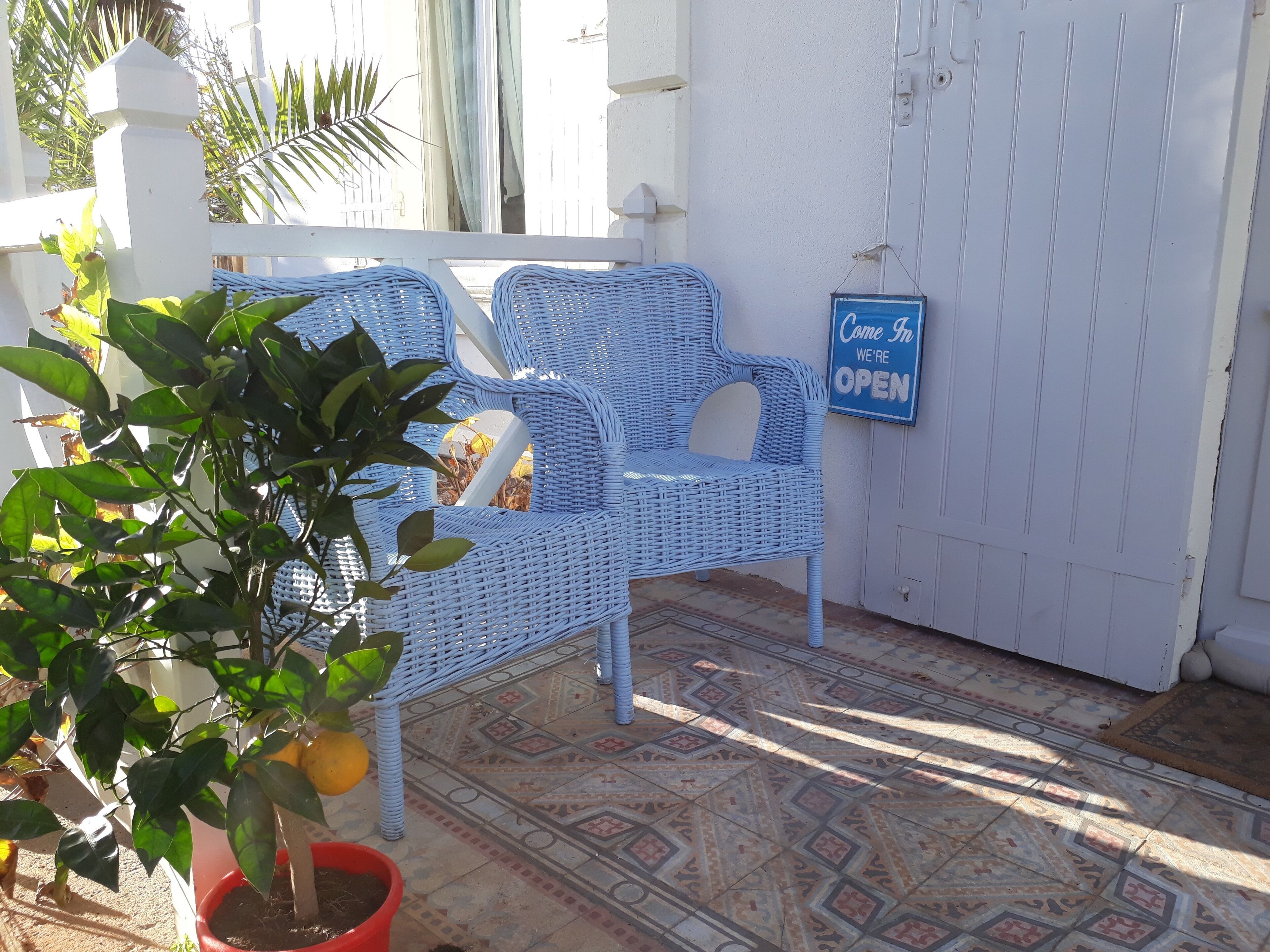 Une chaise en osier bleue avec des coussins, une plante en pot et une pancarte « Entrez, nous sommes ouverts » sur un mur blanc.