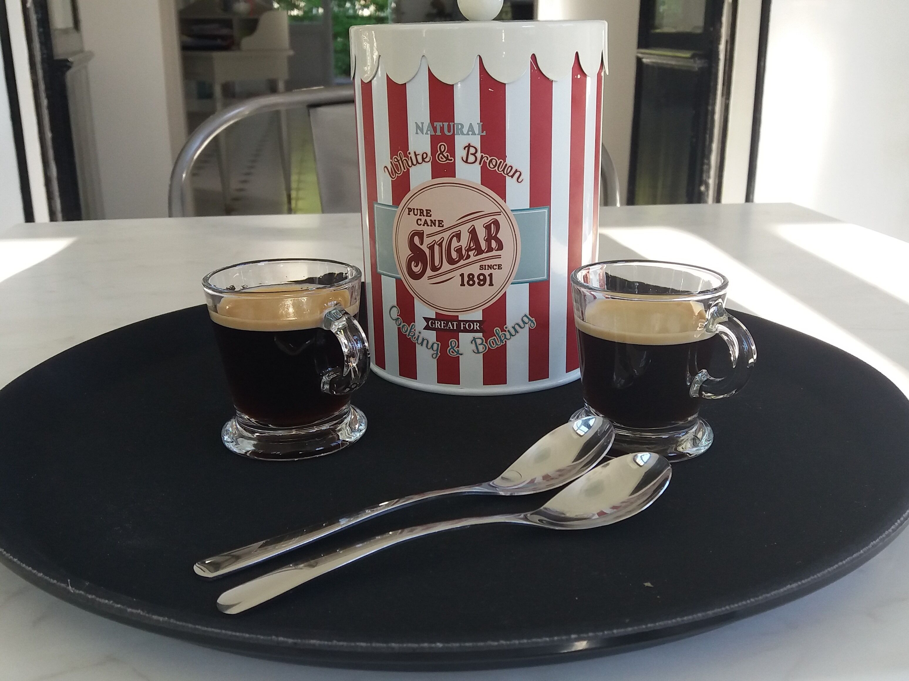 Two shot glasses of dark liquid, a vintage sugar canister, and a spoon on a black tray.