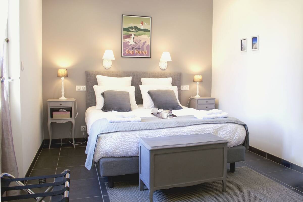 A double bed with two pillows, a bedside table with a lamp, a small bedside table with a lamp, a tray with cups and a teapot, and a framed picture on the wall.