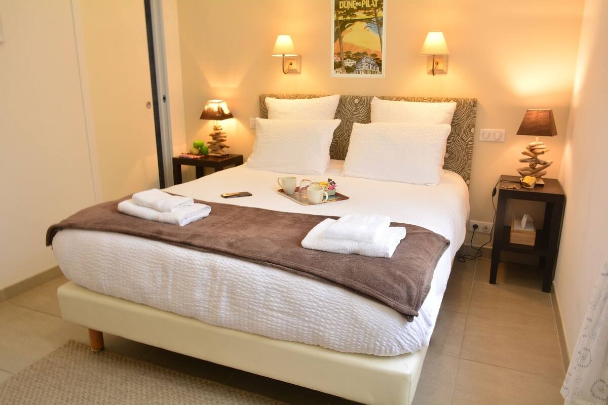 A neatly made bed with white and brown linens, two bedside lamps, a tray with a teapot and cups, and a wall poster.