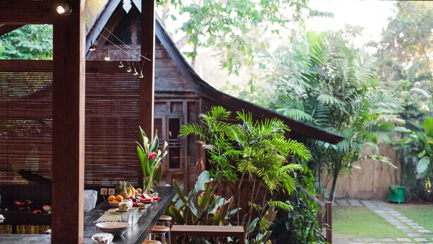 A wooden structure with a dining area featuring a long table, chairs, and a variety of fruits and vegetables.