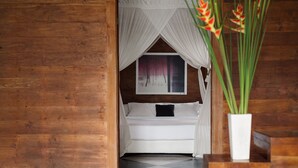 A wooden room with a bed under a white canopy, a painting on the wall, and a potted plant.