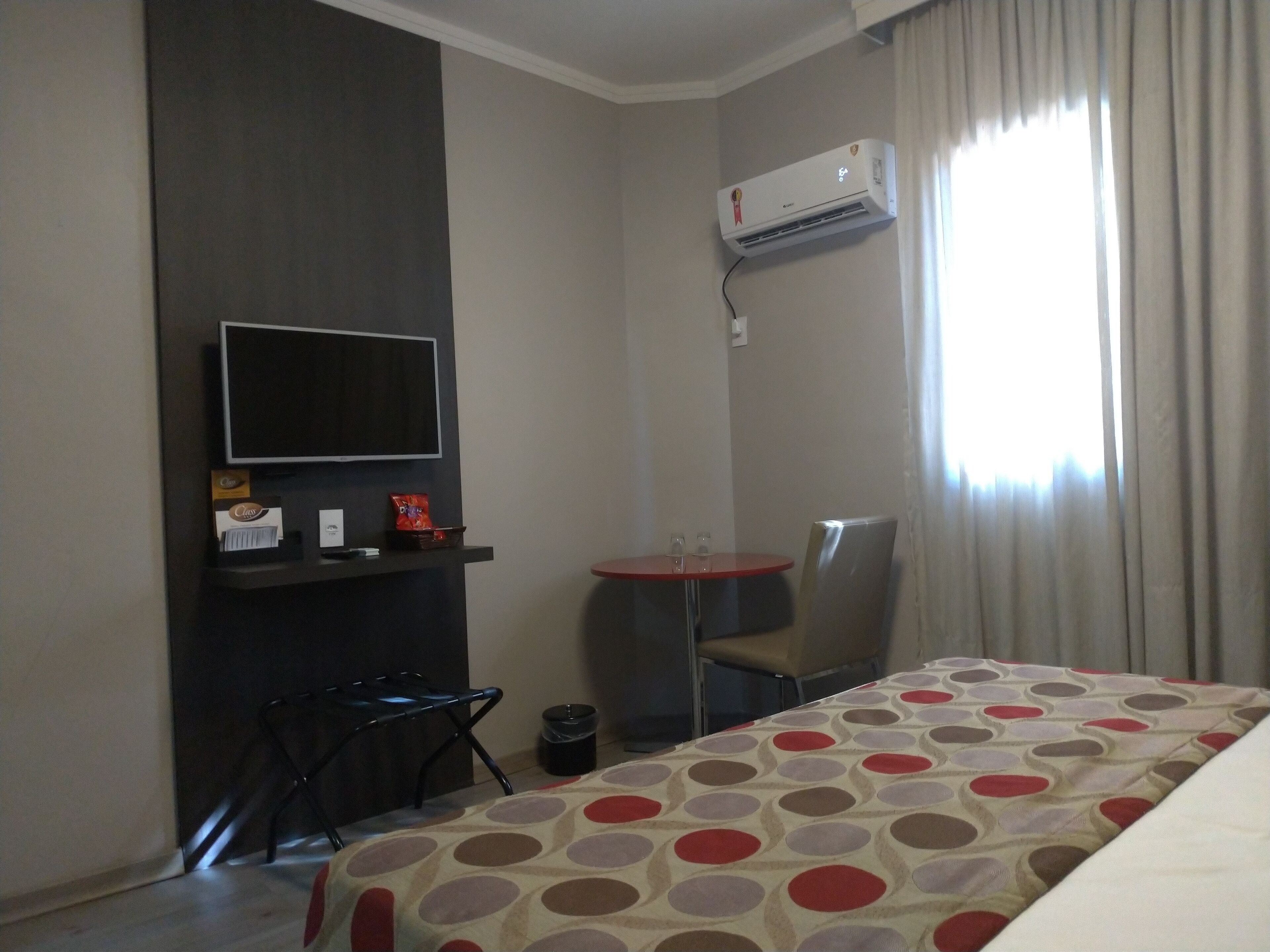 A hotel room with a bed, a TV mounted on the wall, a small table with chairs, and an air conditioning unit.