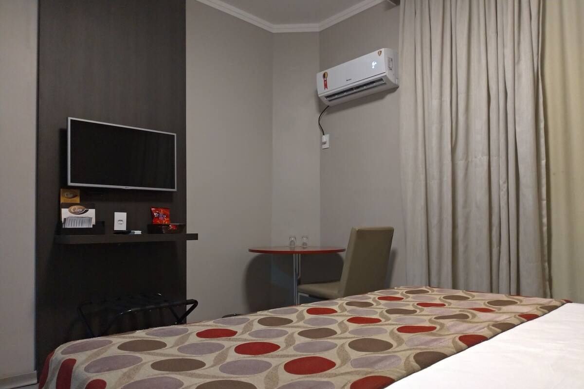 A hotel room with a bed, a TV, a small table, a chair, and an air conditioning unit.