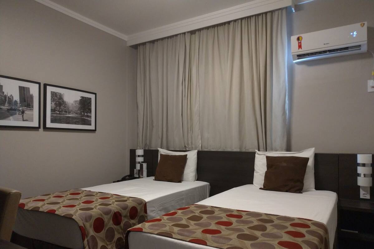 A hotel room with two single beds, a nightstand with a lamp, a wall-mounted air conditioner, and two framed pictures on the wall.