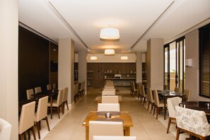A modern dining area with multiple tables and chairs, a central light fixture, and a kitchen in the background.