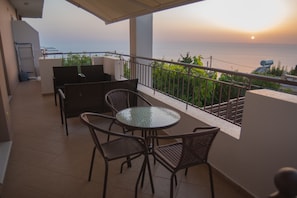 Un balcón con una mesa y sillas de vidrio, con vista a un paisaje marino al atardecer.