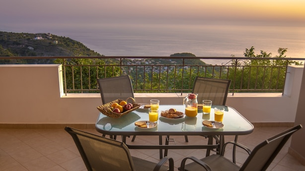 Una mesa dispuesta en un balcón con alimentos para el desayuno, con vistas panorámicas a colinas y al mar.