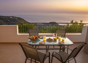 Una mesa dispuesta en un balcón con alimentos para el desayuno, con vistas panorámicas a colinas y al mar.