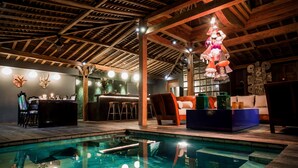 An indoor pool area with wooden beams, a bar, seating, and a hanging light fixture. - Villa Gambar (Kerobokan)