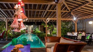 An indoor pool area with wooden beams, hanging lamps, and a dining setup with chairs and tables. - Villa Gambar (Kerobokan)