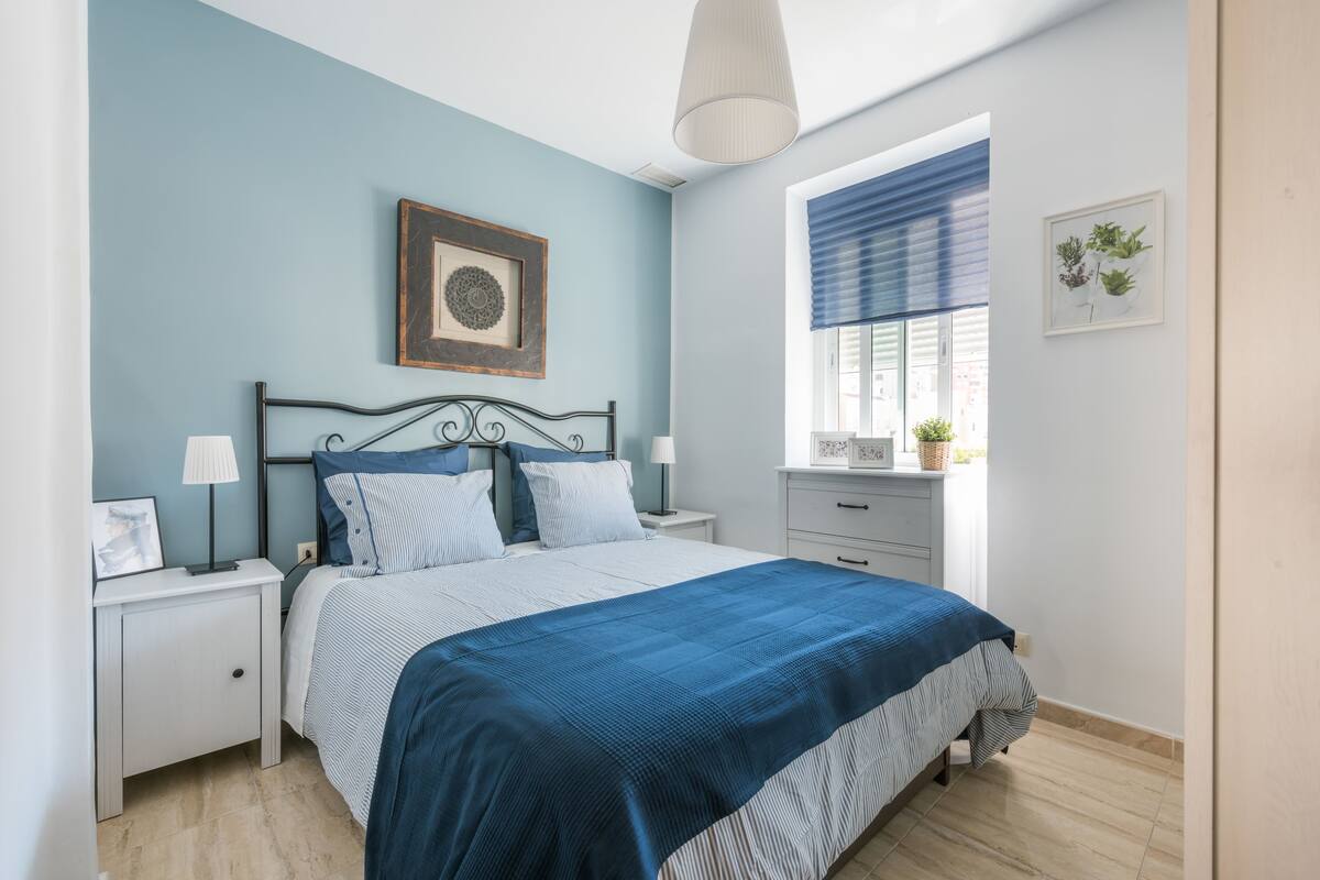 Une chambre à coucher comprenant un lit, des tables de chevet, une fenêtre avec des stores et un tableau accroché au mur.