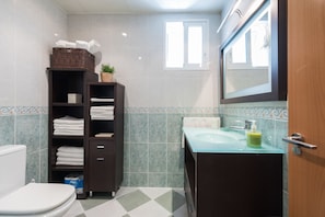 A bathroom with a toilet, a glass sink, a mirror, and a shelf with towels and toiletries.