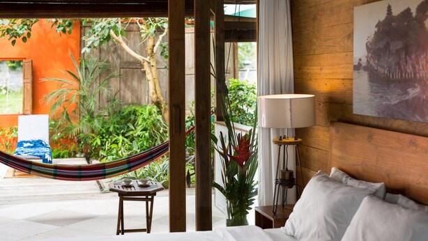A bedroom with a bed, a bedside table, a lamp, and a view of a garden with a hammock. - Villa Madah (Kerobokan)