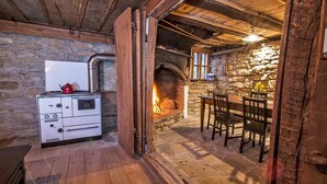 A rustic kitchen with a stone wall, a wood-burning stove, and a dining area with a table and chairs. - Karashka - Vacation Home (Sevlievo)
