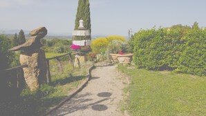 Ein Gartenweg mit einer Steinskulptur, einer hoch aufragenden Zypresse und Blumenbeeten.