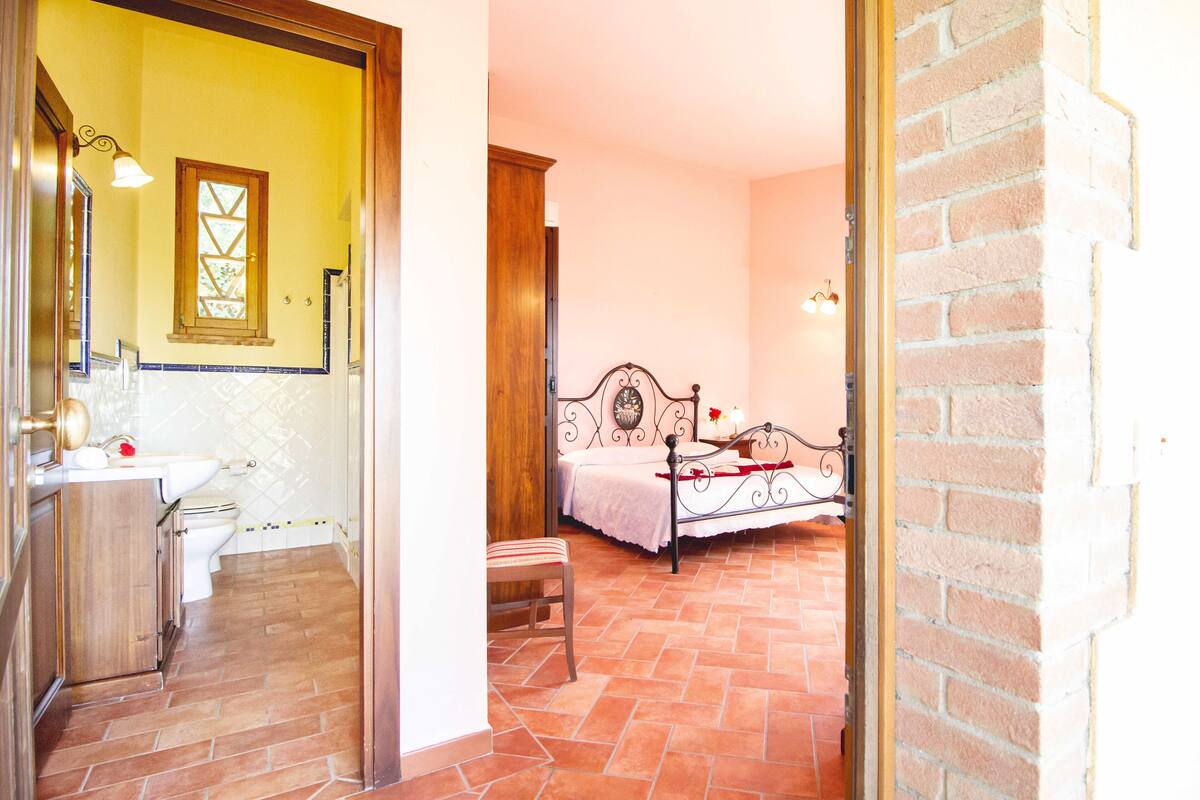 Une pièce comprenant un lit, un petit tabouret, une porte en bois et une salle de bain avec des toilettes et un lavabo.