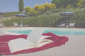 Een zwembadgedeelte met ligbedden en parasols, een boek op een handdoek, en een zwembad op de achtergrond.