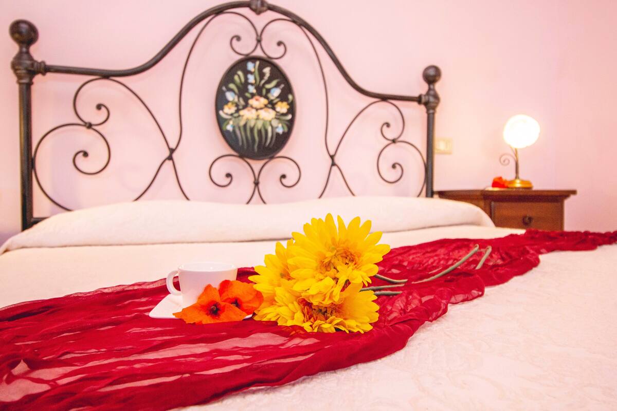 Un lit bien fait, avec un oreiller blanc et un tissu rouge. Sur le lit, on peut voir une tasse blanche et un bouquet de fleurs jaunes.