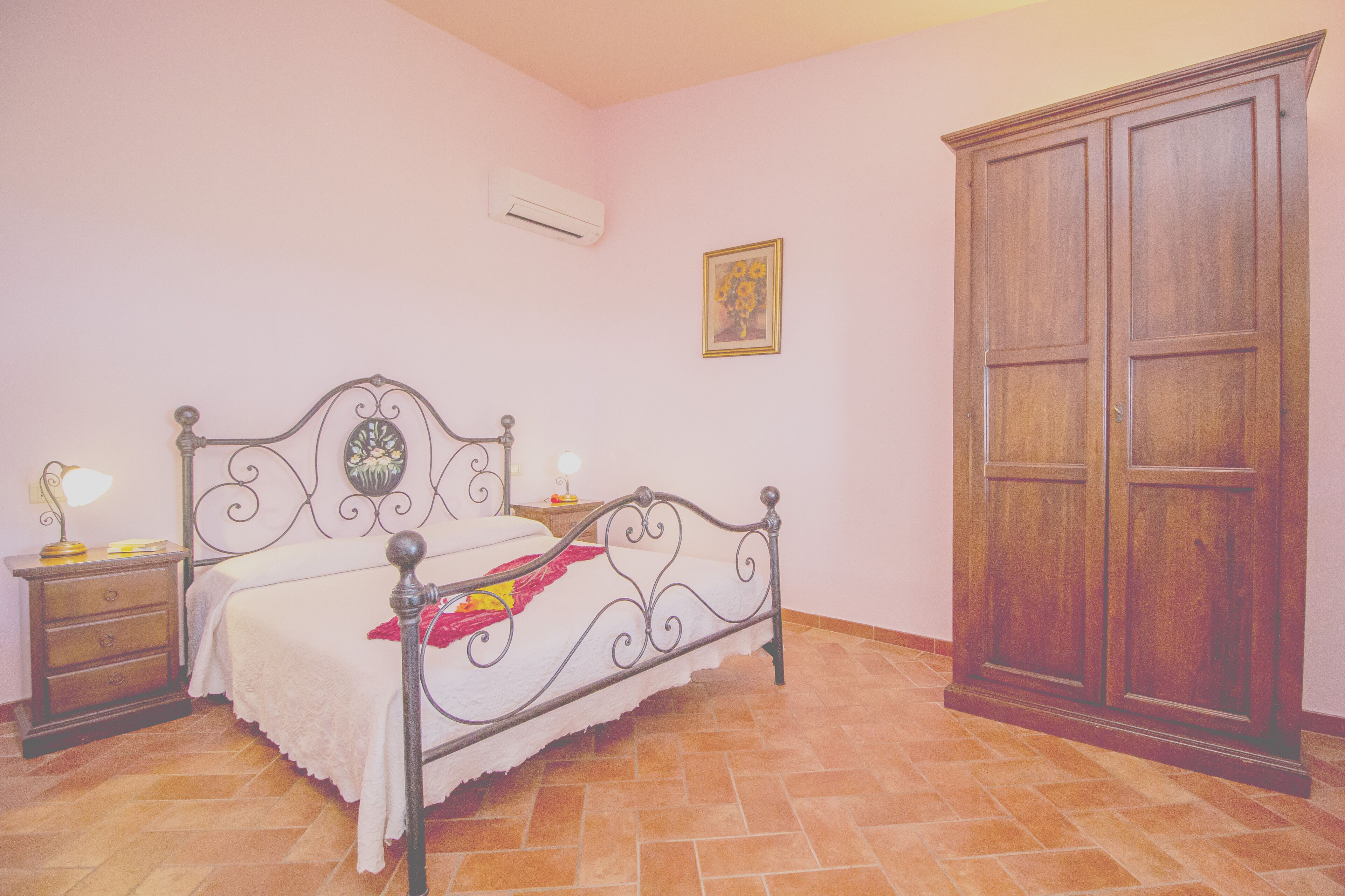 Une chambre à coucher avec un lit à ossature métallique, une armoire en bois, une table de chevet avec une lampe et un climatiseur fixé au mur.