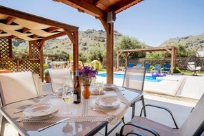 Un área de comedor al aire libre con una mesa dispuesta para dos, con vista a una piscina y colinas.