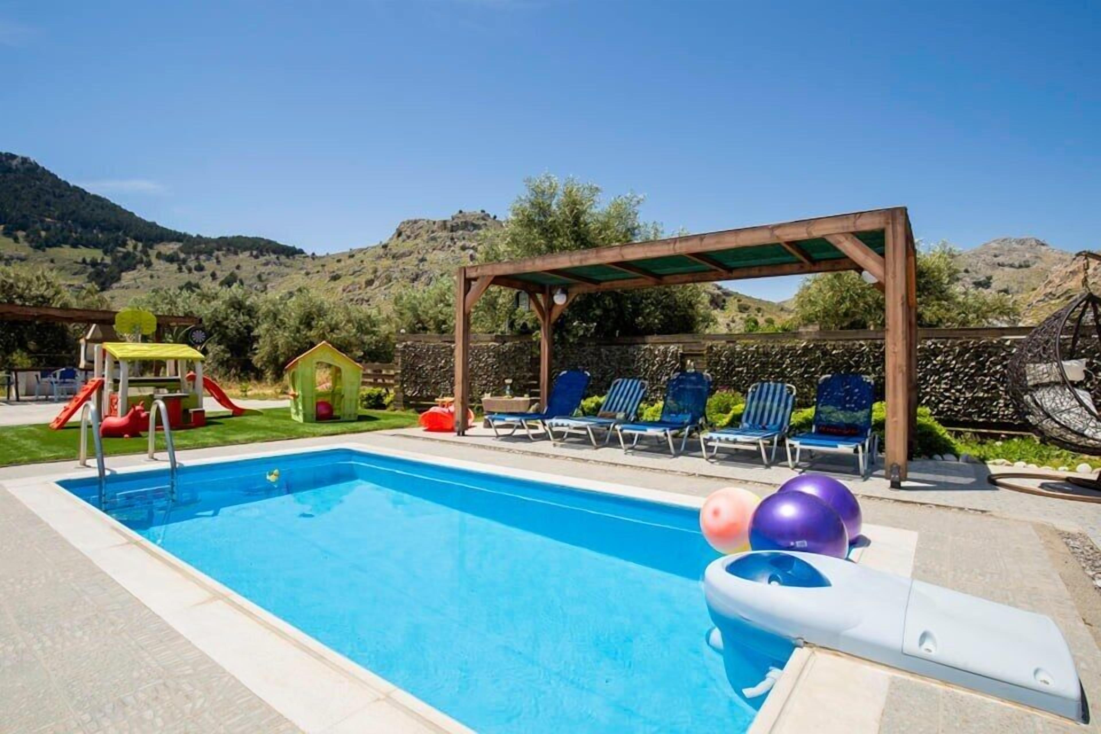 En swimmingpool med et træbeklædt pergola, liggestole og et legeområde for børn.