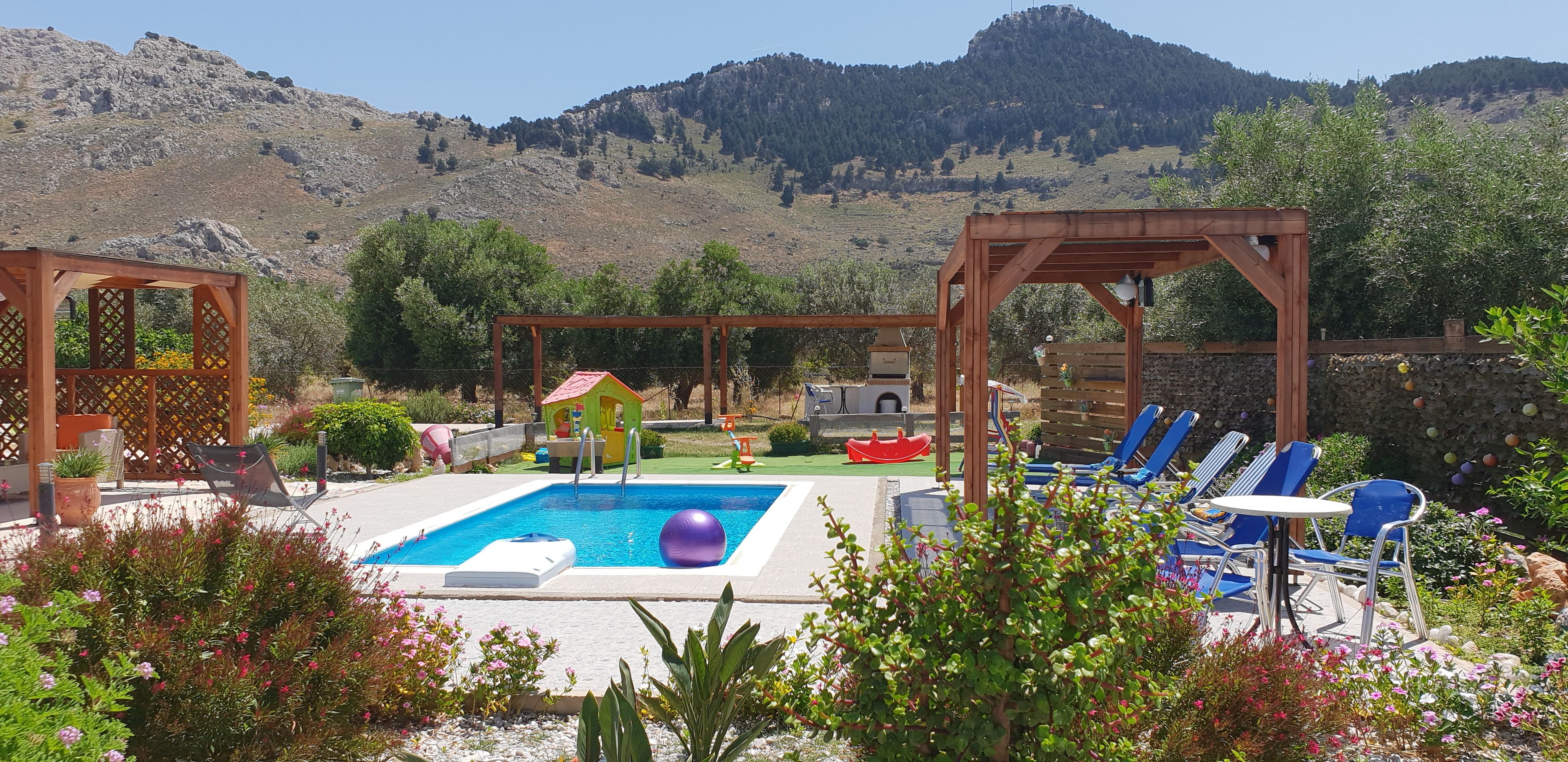 Area piscina con una pergola in legno, poltrone relax e una piccola casetta da gioco.