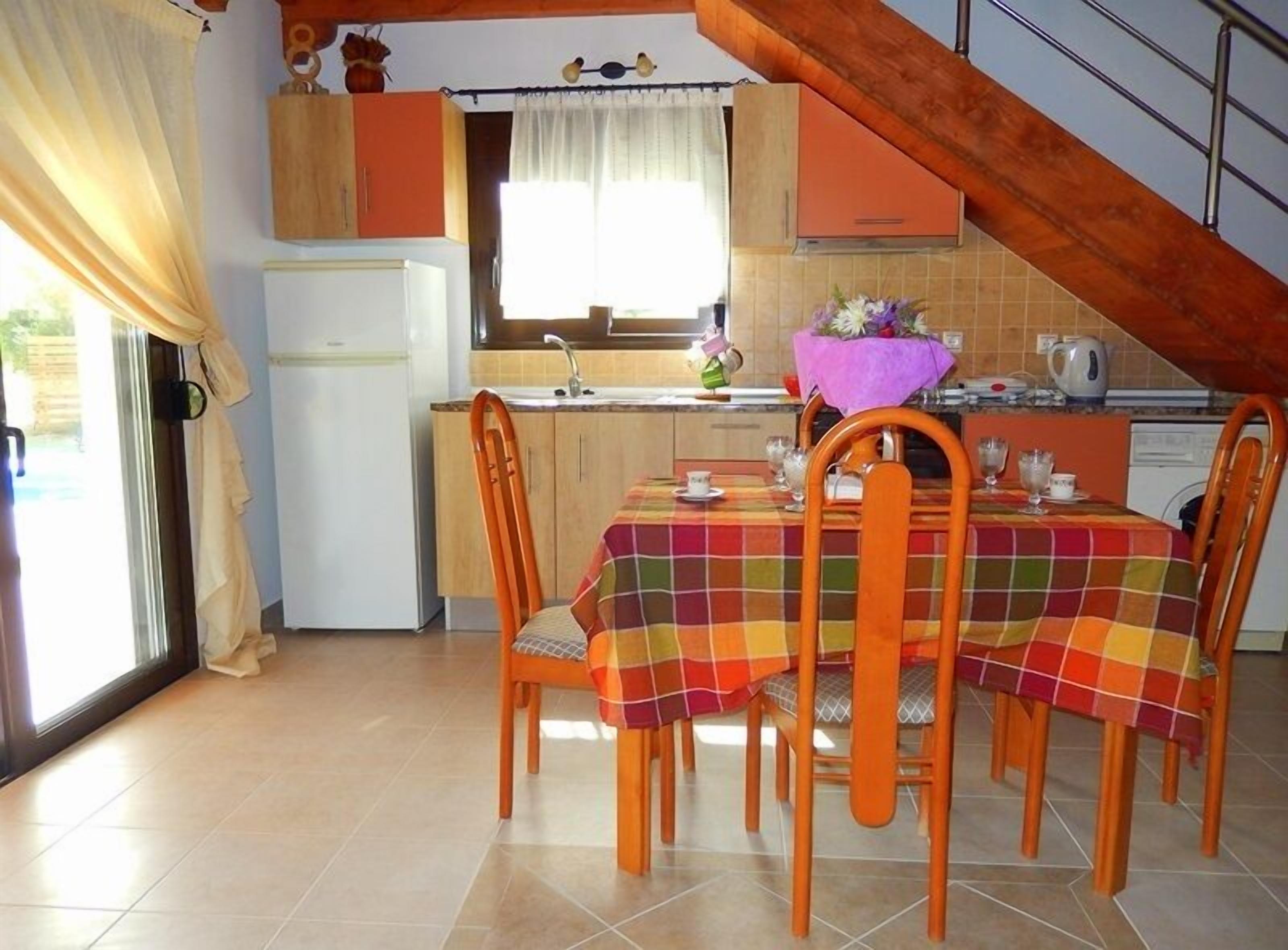 Una cocina con una mesa de comedor preparada para cuatro personas, sillas de madera, un ventanal con cortinas y una escalera.