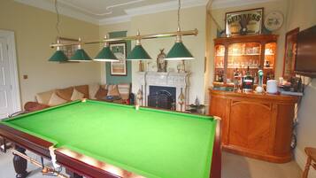 A pool table with a green felt surface, a wooden cabinet with glassware, and a fireplace with a mantel.