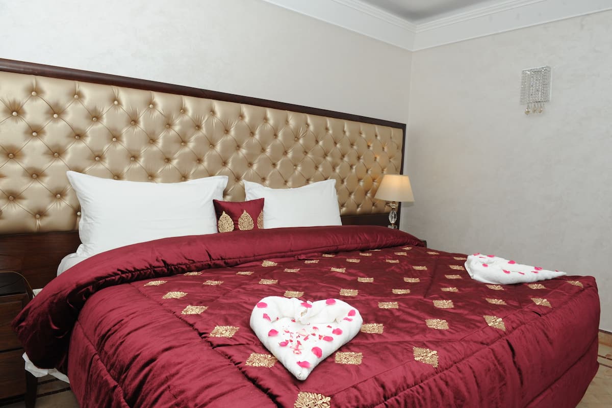 Une chambre d’hôtel avec une tête de lit capitonnée, une couvre-lit rouge et dorée, des oreillers blancs et une décoration en forme de cœur sur le lit.