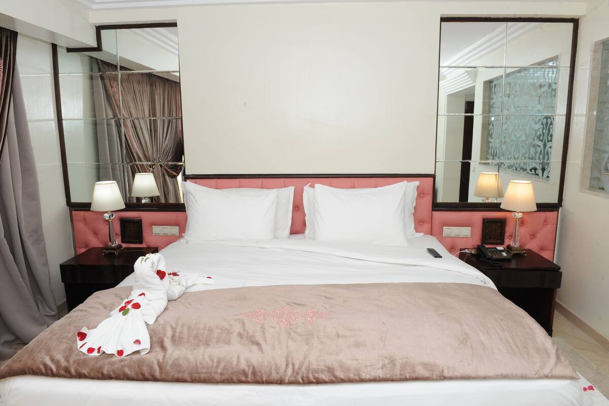 Un lit bien fait, avec du linge de lit blanc, une tête de lit rose et un plaid marron. Il y a deux tables de chevet avec des lampes et un miroir au mur.