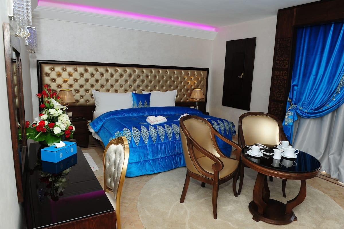 Une chambre d’hôtel avec une tête de lit capitonnée, un lit avec une literie bleue, une chaise en bois, une petite table avec des tasses et un vase de fleurs.