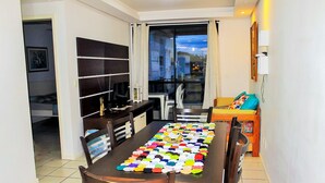 A compact living space with a dining table covered with a colorful table runner, a television on a stand, and a small seating area with a sofa and chairs. - Apartamento Leonardi (Florianópolis)