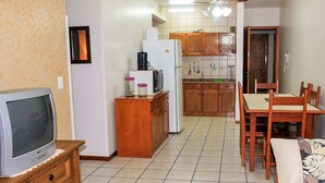 A compact kitchen with a refrigerator, microwave, and wooden cabinets. There is a dining table with chairs and a television on a wooden stand. - Apartamento Leonardi (Florianópolis)
