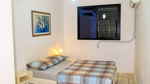 A small, clean room with a bed, a bedside table, a framed picture on the wall, and a window with blinds. - Apartamento Leonardi (Florianópolis)