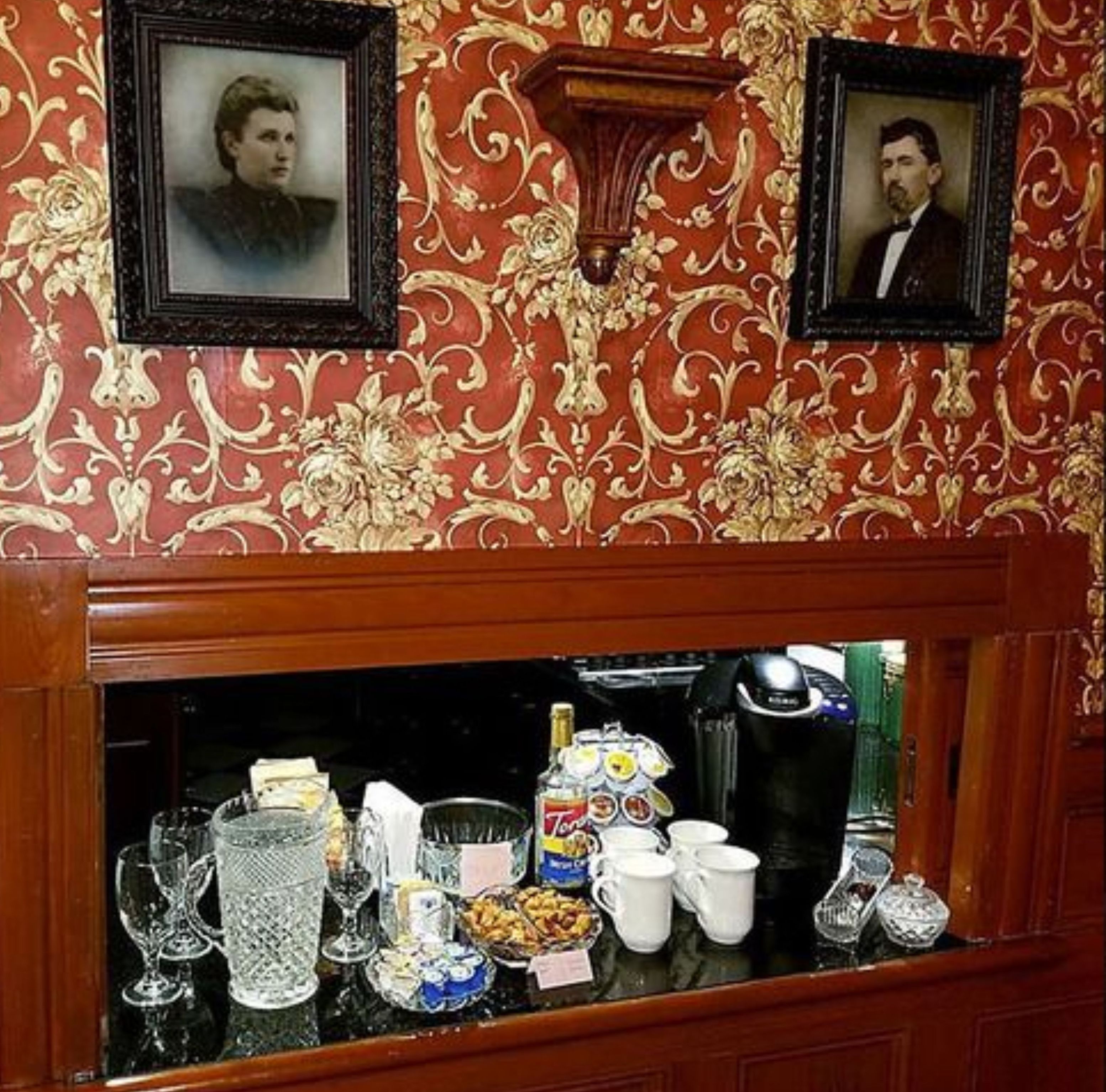 Un espace bar agrémenté d’un papier peint à motifs, de tableaux encadrés accrochés au mur, et d’un assortiment de plats et de boissons présentés sur le comptoir.