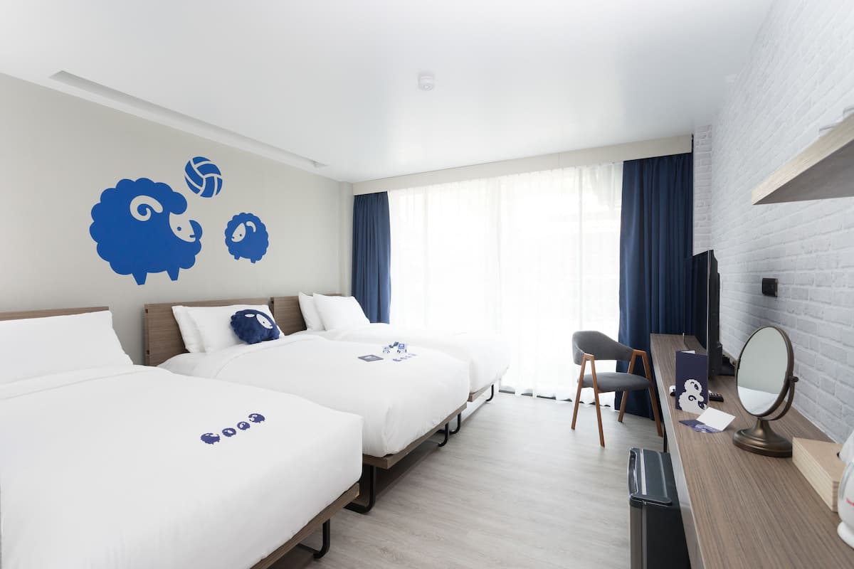 Habitación de hotel con dos camas, un escritorio con silla, un espejo y un ventanal grande con cortinas.