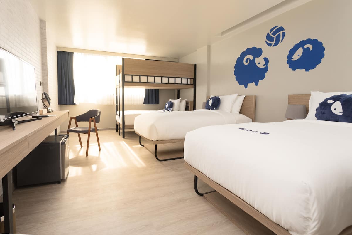 Habitación de hotel con literas, un escritorio con silla, un televisor y una cama con edredón blanco.