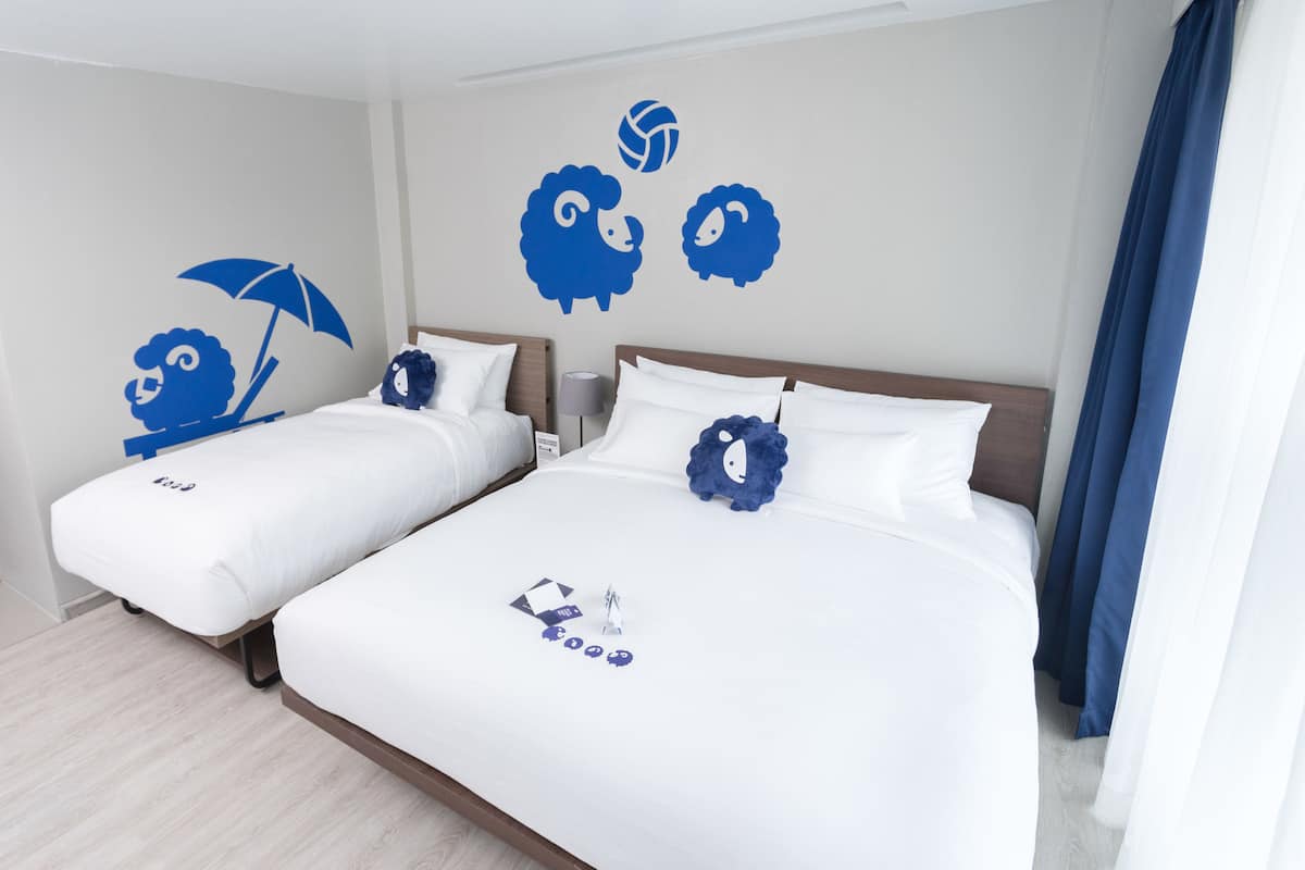 Habitación de hotel con dos camas, ropa de cama blanca y decoraciones azules en la pared, incluyendo ovejitas y una pelota de voleibol.