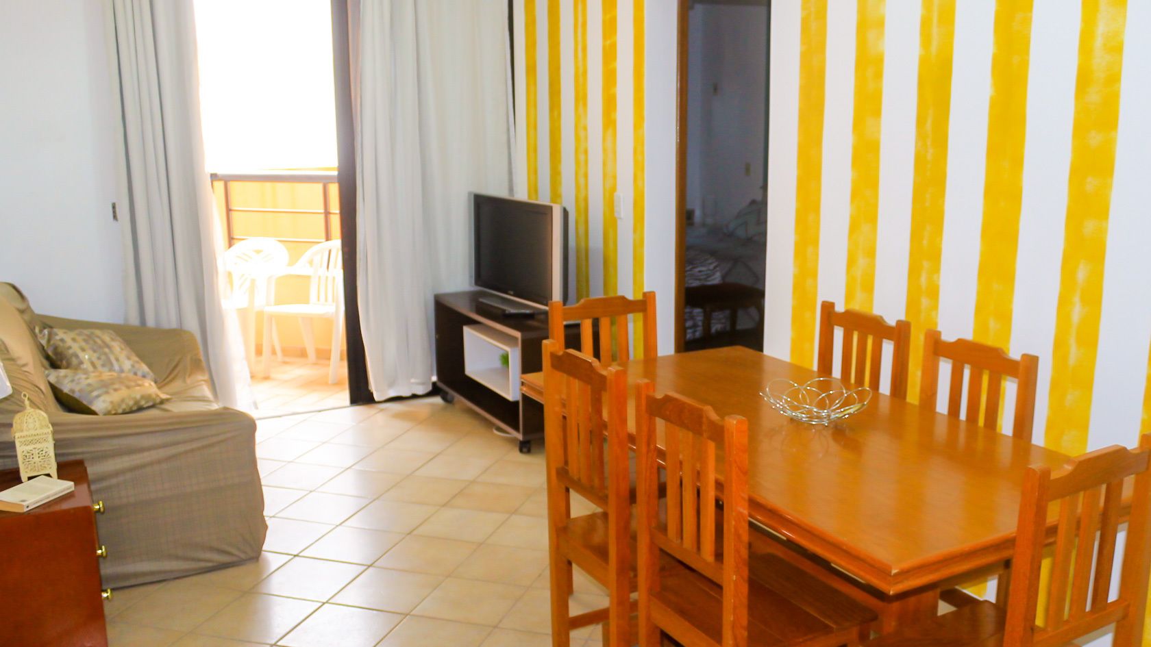 A living room with a sofa, a TV, a dining table with chairs, and a kitchen area with yellow-striped walls.