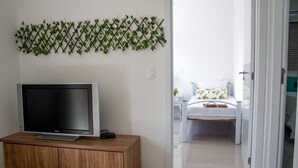 Un dormitori modern amb un televisor sobre un moble de fusta, un llit amb roba de llit blanca i una decoració de paret amb fulles de vitalàries.
