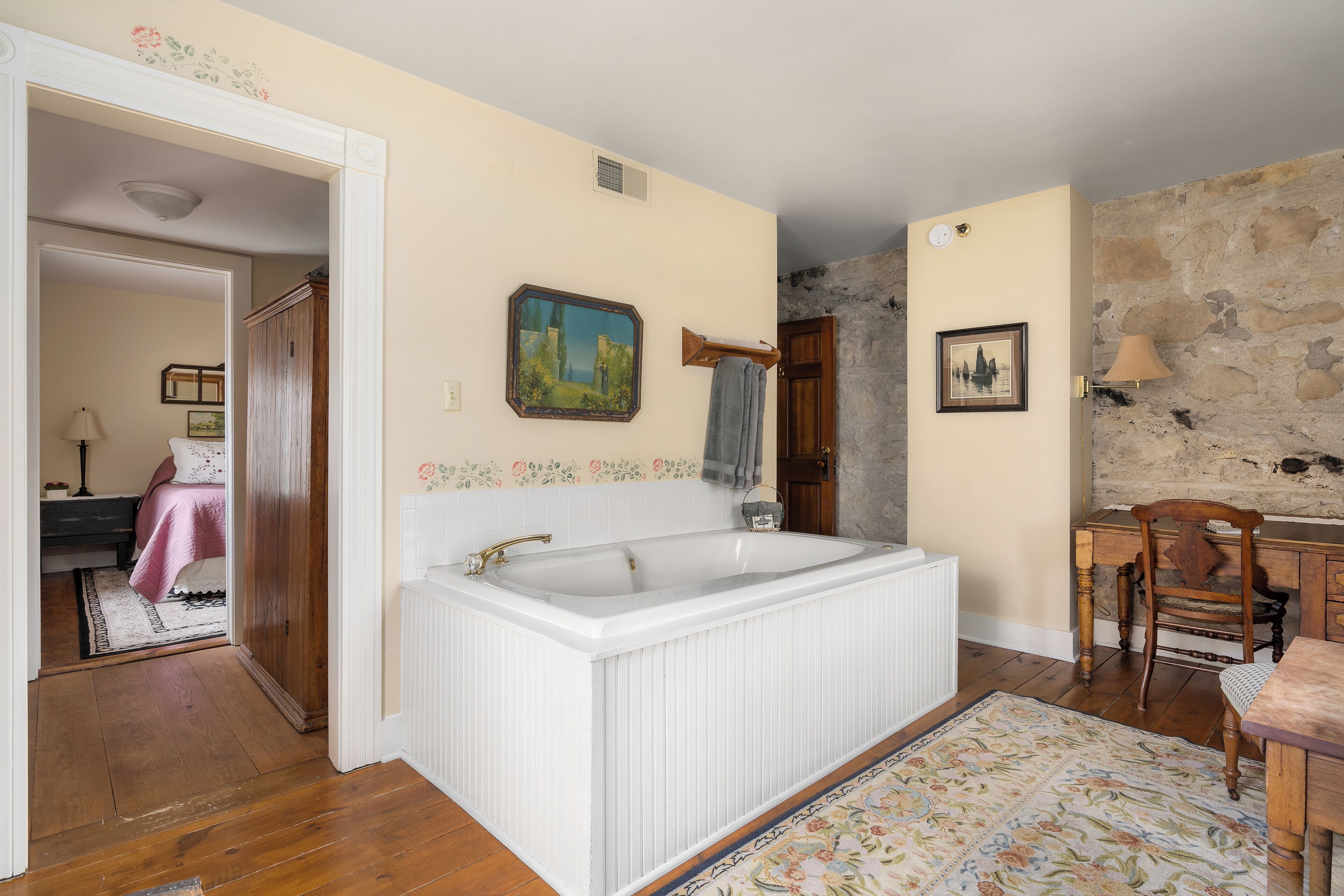 A room with a large white bathtub, wooden furniture, a stone wall, and a door leading to another room.