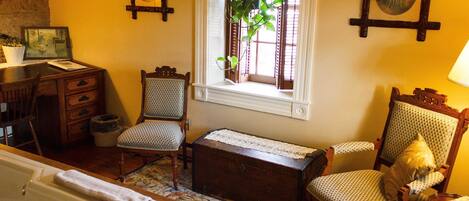 A traditional room with a wooden desk, two upholstered chairs, a wooden chest, a window with a plant, and a framed portrait on the wall.
