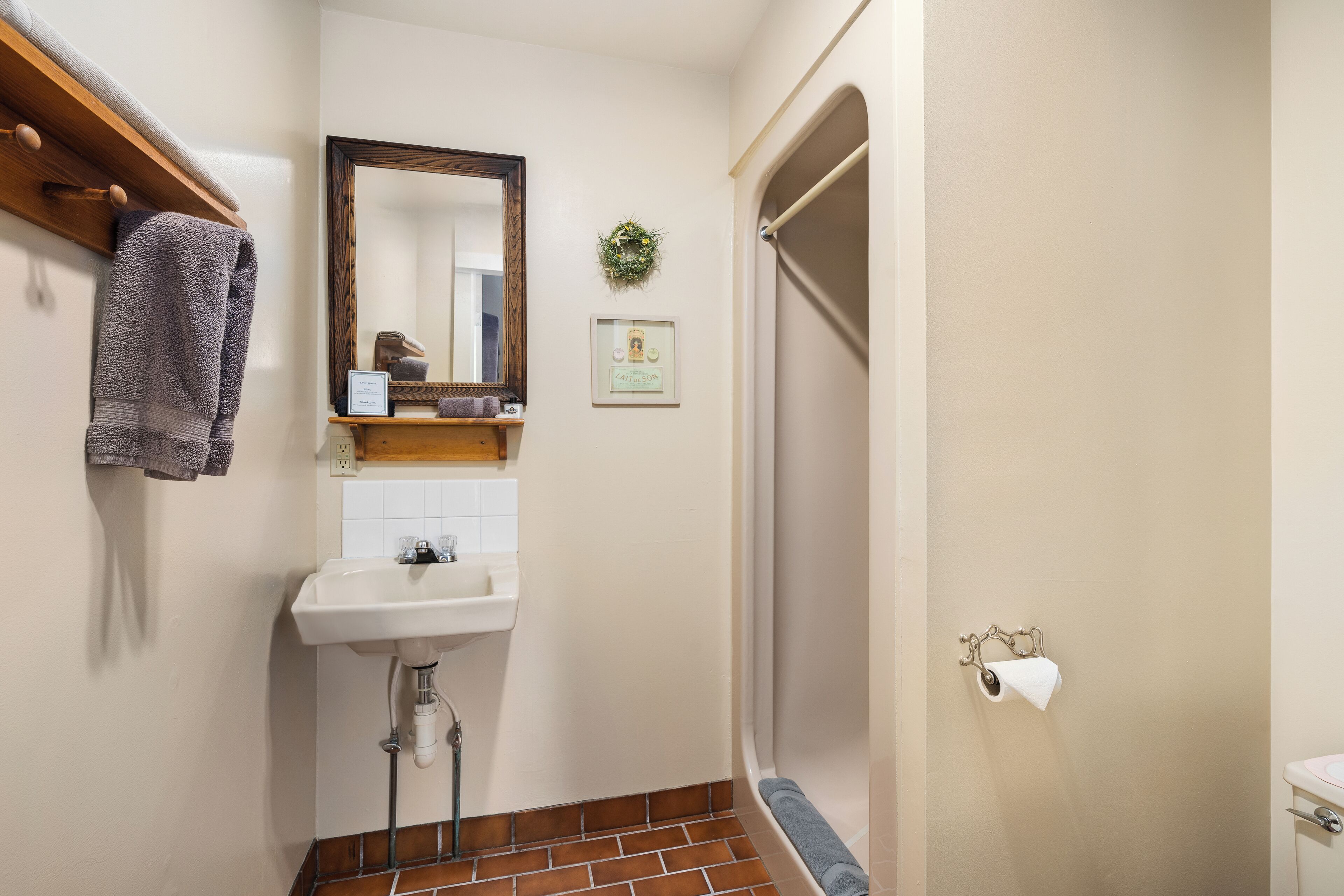Un baño con lavamanos, espejo, toallero, ducha y dispensador de papel higiénico.