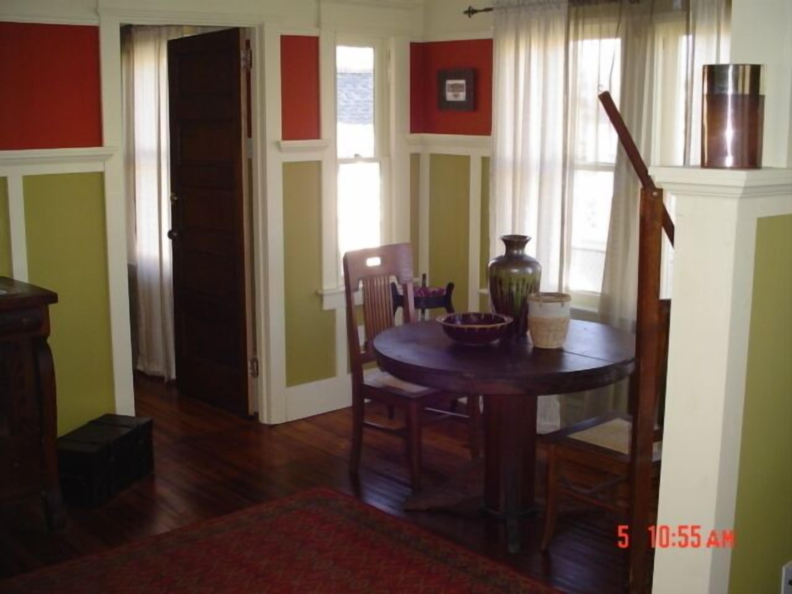 A dining room with a round table, chairs, and a vase. There is a door and a window with curtains.