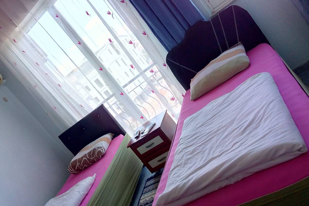 A room with a bed, a window with curtains, a wall-mounted air conditioner, and a small table with a chair.