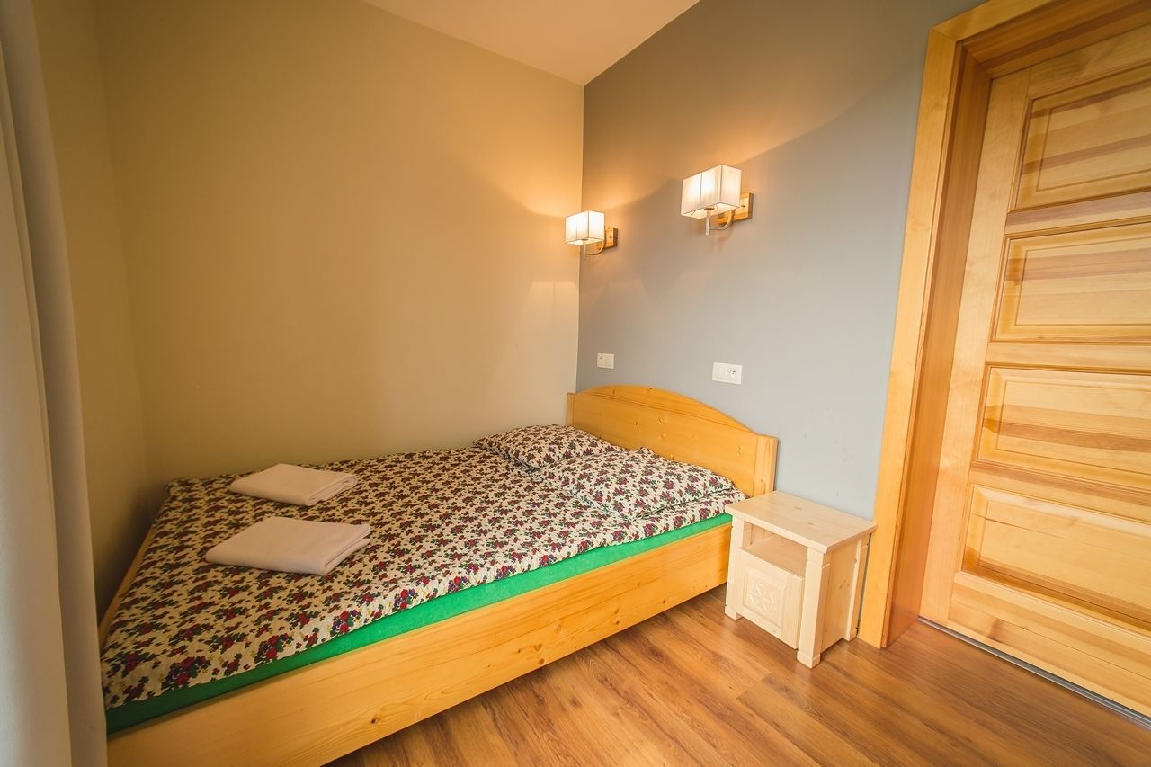 Un lit simple avec une couvre-lit à motifs floraux, deux lampes fixées au mur, une tête de lit en bois, une petite table de chevet et une armoire en bois.